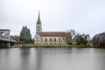 A long-exposure photograph capturing the imposing, Gothic architecture of a riverside church, complete with a tall spire, situated directly on the bank of a smooth, calm river, with a graveyard