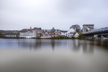 A long-exposure photograph across a very smooth, reflective river showing a row of grand, traditional buildings, including a large white structure and red-brick houses , marlow