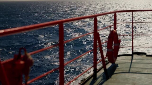 Sunlit ocean waves shimmer beyond a red ship deck railing as a vessel journeys toward a distant horizon, revealing tranquil water and the vast openness of the sea.