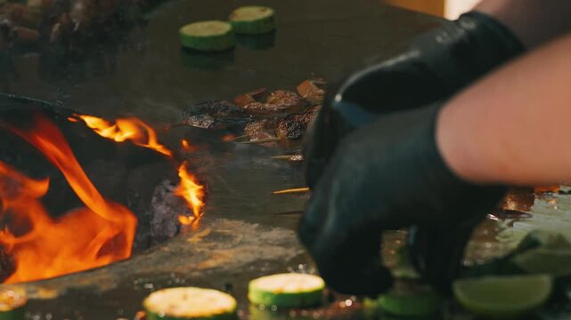 A close-up of sizzling shrimp skewers and grilled zucchini, the surface licked by flames. A gloved hand places the zucchini on the hot surface, creating a delightful cooking atmosphere