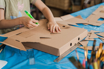 Tropical Christmas DIY art class with kids and diverse women workshop leaders making an eco-friendly cardboard holiday house, visible greenery and warm weather outside, boy wearing Santa hat proudly