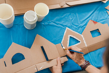 Tropical Christmas DIY art class with kids and diverse women workshop leaders making an eco-friendly cardboard holiday house, visible greenery and warm weather outside, boy wearing Santa hat proudly
