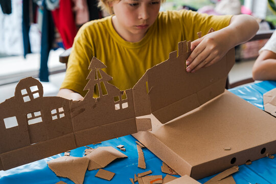 Tropical Christmas DIY art class with kids and diverse women workshop leaders making an eco-friendly cardboard holiday house, visible greenery and warm weather outside, boy wearing Santa hat proudly