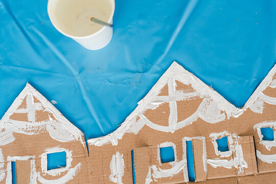 Tropical Christmas DIY art class with kids and diverse women workshop leaders making an eco-friendly cardboard holiday house, visible greenery and warm weather outside, boy wearing Santa hat proudly