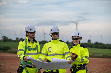 Engineers wearing safety helmets inspecting a wind turbine at a wind farm and holding a blueprint while working.