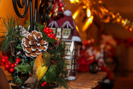 A detailed close-up of Christmas ornament featuring a small decorative bird, pinecone, and red berries, with a miniature festive house and warm string lights blurred in the background