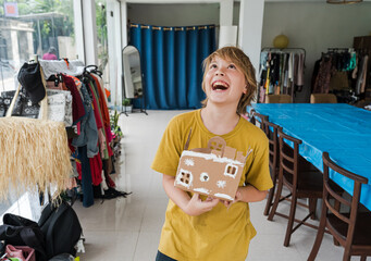 Tropical Christmas DIY art class with kids and diverse women workshop leaders making an eco-friendly cardboard holiday house, visible greenery and warm weather outside, boy wearing Santa hat proudly