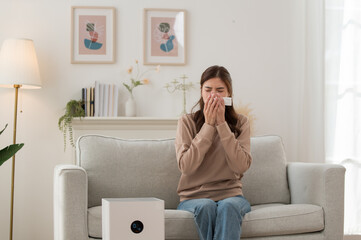 Asian woman sneezing and using tissue while sitting on sofa at home with air purifier