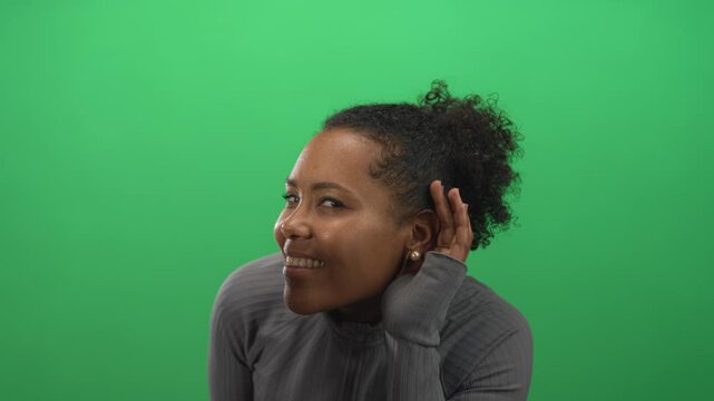 Young african american woman hand to ear for listening while smiling on green studio set; curiosity.