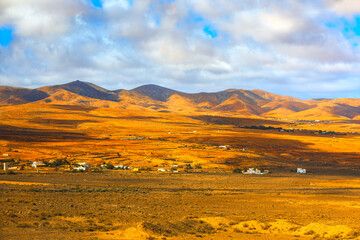 Arid desert landscape of Valle de Santa Ines in Fuerteventura, Spain. View includes dry mountains, a small settlement of white buildings, and a cloudy sky