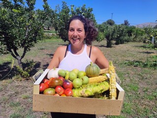 Smiling Woman Holding a Box of Fresh Organic Vegetables in a Sunny Orchard