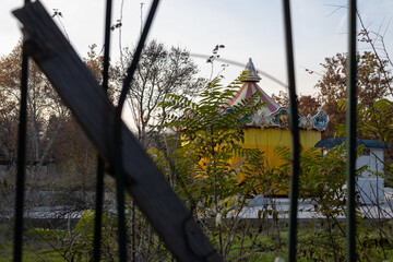 an abandoned, colorful carousel building is viewed through dark, vertical slats and overgrown, dry foliage on a gloomy, late autumn afternoon
