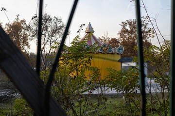 an abandoned, colorful carousel building is viewed through dark, vertical slats and overgrown, dry foliage on a gloomy, late autumn afternoon