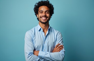 Obraz premium Handsome Hispanic man smiles confidently against vibrant blue backdrop. Wears light blue shirt, crossed arms. Portrait conveys positivity, self-assurance, suitable for diverse business contexts.