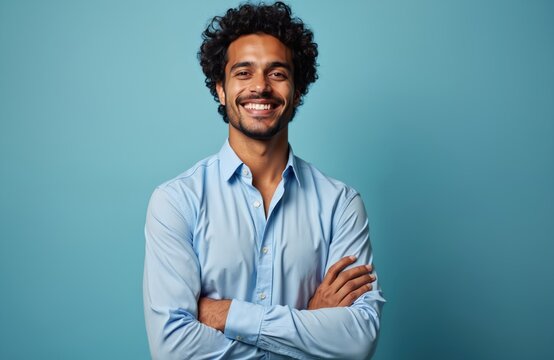 Handsome Hispanic man smiles confidently against vibrant blue backdrop. Wears light blue shirt, crossed arms. Portrait conveys positivity, self-assurance, suitable for diverse business contexts.