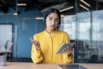 Portrait of a young Indian woman in a headset holding a tablet, sitting at a table and talking to the camera on a video call