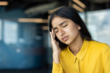Close-up photo of a young Indian woman in an office, holding her head in her hand and grimacing in pain