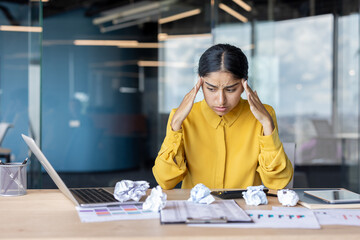 A sad young Indian woman sits frustrated at an office desk with crumpled paper and holds her head...