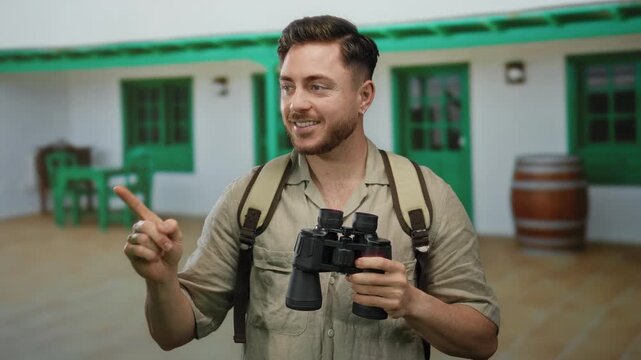 Young tourist man with a beard, holding binoculars and pointing on a city street, wears a backpack, suggesting adventure and exploration.