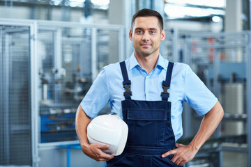 Confident industrial mechanic in blue overalls with helmet in a factory.