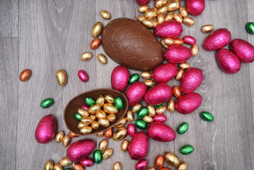 Group of colourful foil wrapped chocolate easter eggs in pink, red, gold and green. Large halves of a brown milk chocolate egg have mini eggs inside, on a grey wooden background.