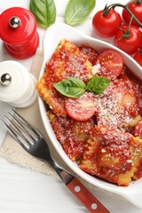 Delicious ravioli with tomato sauce, cheese and basil in baking dish served on white wooden table, flat lay