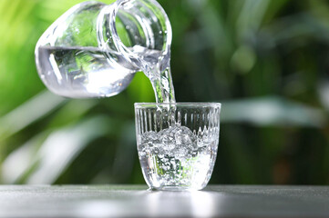 Pouring water into glass from jug at grey table against blurred green background, closeup. Space for text