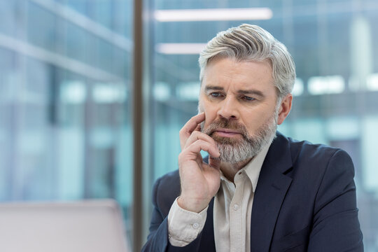 Mature bearded businessman looking at a laptop screen, concentrating on a serious issue, reflecting on a challenge, and finding a solution in a modern office environment - Powered by Adobe