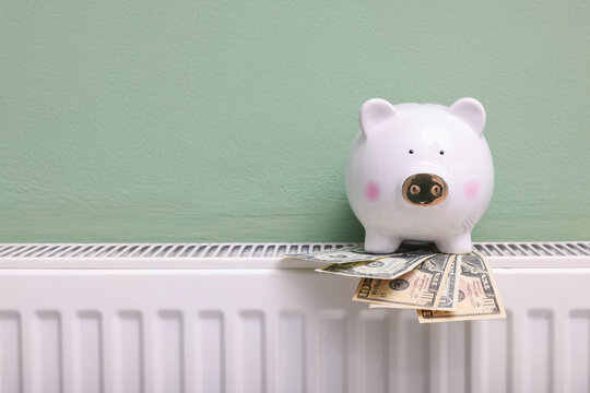 Piggy bank and dollar banknotes on heating radiator near green wall, closeup - Powered by Adobe