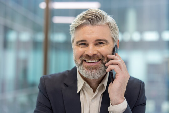 Confident mature businessman with a beard smiling while talking on a mobile phone, representing professional communication, success, and leadership in a modern office environment