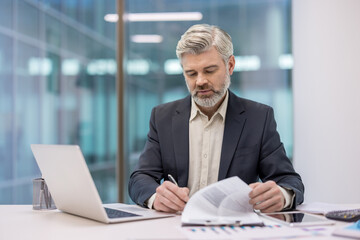 Mature businessman with gray hair and beard signing an official contract, reviewing important legal documents while working at his desk in a modern corporate office setting