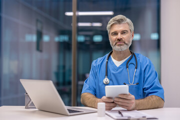 Experienced male doctor wearing scrubs and a stethoscope, looking at the camera while holding a digital tablet in a modern medical office, representing healthcare technology and digital medicine