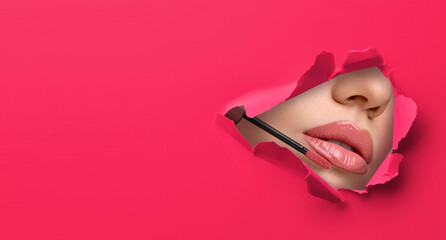 Young woman applying lip gloss, closeup. View on lips through torn hole in bright pink paper