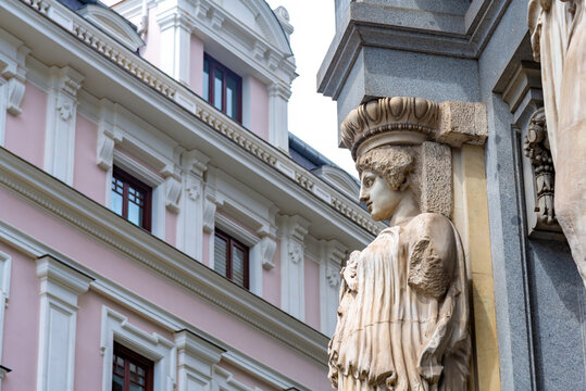 A close-up of a classical female statue carved into the Cervantes Institute facade, set against a blurred pink-and-white building with dark windows under a bright sky
