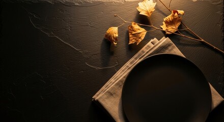 A dark table setting for Thanksgiving. A round black plate rests on a folded gray napkin. Dried autumn leaves and a twig are arranged nearby.