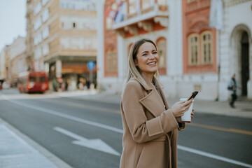 Fototapeta premium Young woman smiling and holding a coffee cup on a lively city street in autumn