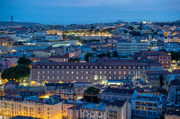 Obraz premium Illuminated buildings of Lisbon, Portugal in deep afternoon twilight with Tagus River and bridge in background from Miradouro da Senhora do Monte.