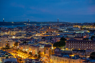Illuminated buildings of Lisbon, Portugal in deep afternoon twilight with Tagus River and bridge in background from Miradouro da Senhora do Monte.