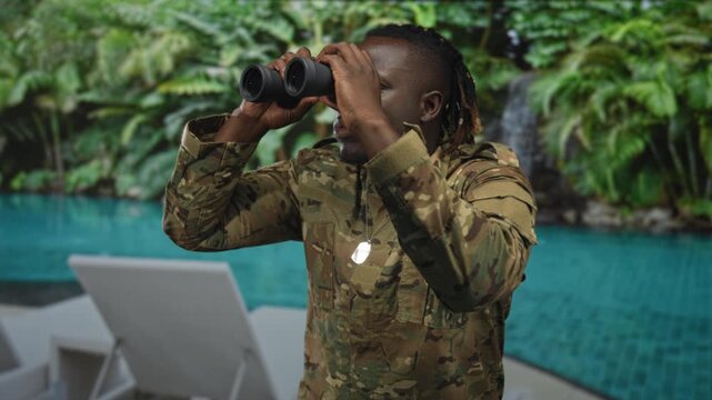 Man soldier in camouflage uniform looking through binoculars at resort pool with lounge chairs and waterfall, dog tags visible; duty vigilance.