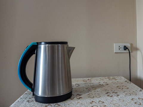 A sleek, brushed stainless steel electric kettle with a vibrant teal handle sits on a subtly patterned tablecloth, conveniently plugged into a wall outlet. This modern appliance is ready to boil water