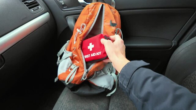 hand retrieves red first aid kit from orange backpack on passenger seat, driver sleeve visible, car interior grey dashboard, roadside stop, urgent calm response, hiking gear ready for emergency