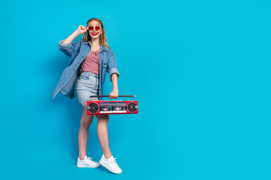 Glamorous young woman in denim jacket and red sunglass with boombox against blue background for fashion lifestyle and shopping promotion
