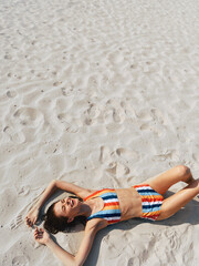 Fototapeta premium Young woman lying on white sand beach in a colorful striped bikini, sunlit and relaxed, capturing a carefree summer moment with warm tones and gentle breeze.