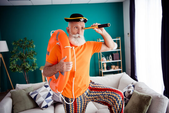 Funny retro pirate at home explores a bright living room wearing an orange shirt zigzag pants and a life jacket while using a telescope for a playful indoor vacation vibe