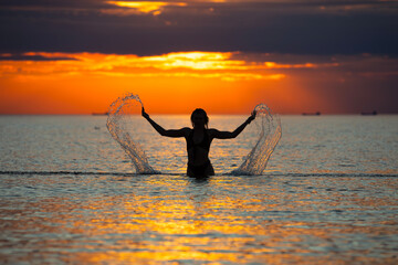 Silhouette of young woman splashing water on the beach at sunset