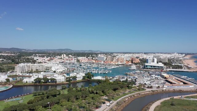 Vilamoura aerial view aerial view vilamoura marina riverfront, bright blue sky and turquoise basin dotted with yachts, white Mediterranean buildings, tree-lined promenade and winding river