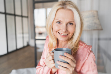 Close up of smiling mature woman in pajamas sitting on comfortable bed with coffee cup at home looking at camera. Middle age lady having morning hot tea while lying on cozy bed in the bedroom