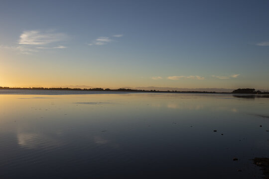 View of the tranquil waters reflecting the gradient sky at dusk, with distant trees lining the horizon, painting a serene picture of nature, Christchurch, Canterbury Region, New Zealand.