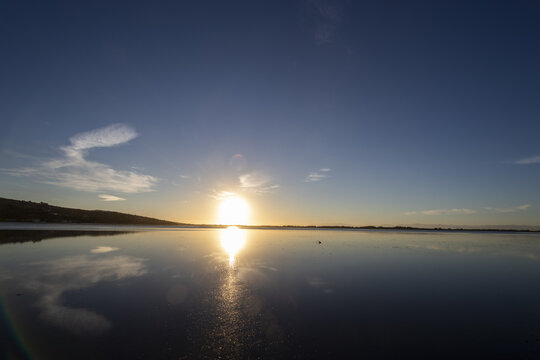 View of the sun's golden orb ablaze on the serene water, mirroring the blue sky above, with a touch of land on the horizon, Christchurch, Canterbury Region, New Zealand.