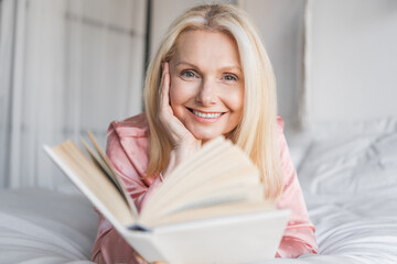 Middle age woman dressed in pajamas sitting comfortably on bed reading a book in cozy bedroom. Natural light coming through window illuminates scene, creating calm atmosphere